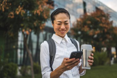 Olgun Japon kadın cep telefonu kullanıyor ve modern binanın önünde duruyor.