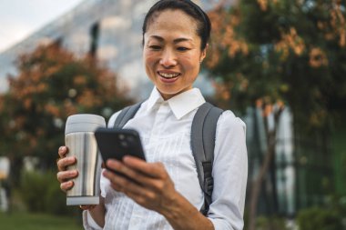 Olgun Japon kadın cep telefonu kullanıyor ve modern binanın önünde duruyor.