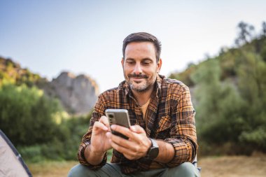 Yetişkin beyaz bir adam kamp yaparken çadırın önüne oturur ve cep telefonunu kullanır.