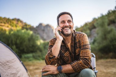 Yetişkin beyaz adam kamp gezisinde çadırın önünde oturur ve cep telefonuyla konuşur.