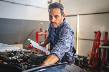 Bir araba tamircisi, bir otomobil atölyesinde elinde bir pano tutarak bir motoru incelemeye odaklanır. İyi ışıklandırılmış sahne onun konsantrasyonunu ve uzmanlığını vurguluyor.