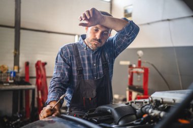 Yorgun araba tamircisi alnını sildi, garajda bir araca yaslandı, modern bir atölyede aletler ve ekipmanlarla çevrili, yorgunluk ve adanmışlık taşıdı.