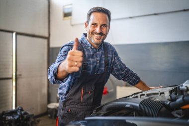 Kendinden emin bir araba tamircisi gülümseyip, bir araba motoruna yaslanırken, becerisini ve çalışmalarının kalitesini iyi aydınlatılmış bir atölyede sergiliyor.