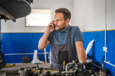 Tulum giymiş özenli bir araba tamircisi, cep telefonuyla konuşurken bir motoru ayarlıyor, sarı ve mavi bir garaj içerisine karşı ayarlanıyor. Odaklanmış ve adanmış çalışma ortamı