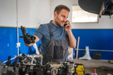 Mavi gömlekli bir tamirci, mavi ve beyaz bir otomobil tamir atölyesi ortamında çalışan bir motor üzerinde çalışırken cep telefonuyla konuşuyor.