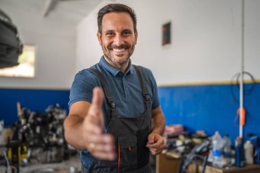 Gülümseyen tamirci dostça bir tokalaşma sergiliyor, bir oto tamir atölyesinde dururken, hoş karşılanan ve ulaşılabilir bir atmosfer yaratıyor.