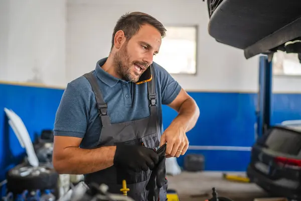 stock image An attentive car mechanic, dressed in overalls, adjusts an engine while talking on the mobile phone, set against a yellow and blue garage interior. Focused and dedicated work atmosphere