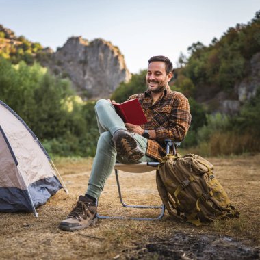 Yetişkin beyaz bir adam çadırın önünde oturur ve kamp gezisinde kitap okur.
