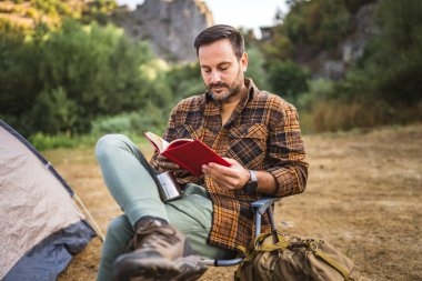 Yetişkin beyaz bir adam çadırın önünde oturur ve kamp gezisinde kitap okur.