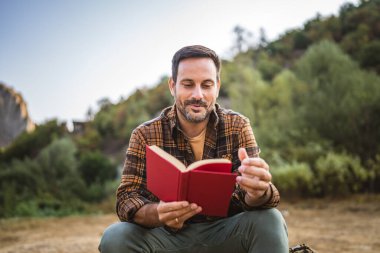 Yetişkin beyaz bir adam çadırın önünde oturur ve kamp gezisinde kitap okur.