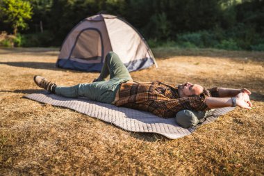 Yetişkin bir kampçı, güneşli bir günde arka planda çadırıyla kuru çimenlerin üzerinde yastıklı bir paspasın üzerinde uzanıyor.