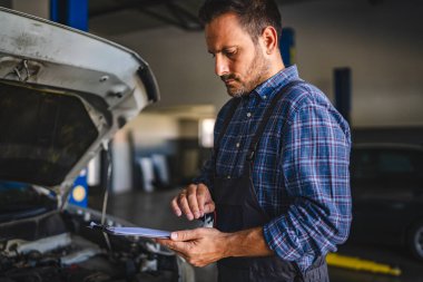 Otomobil tamirhanesindeki bir tamirci açık motor bölümünü inceliyor ve bir pano üzerindeki teşhis sonuçlarını inceliyor. Bu da özenli araç bakımı ve hizmet verimliliğini gösteriyor.