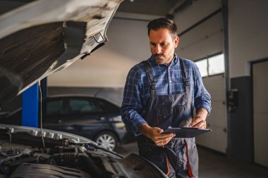 Odaklanmış yetişkin bir araba tamircisi arabanın motorunu inceler, parlak ışıklandırılmış bir atölyede bir pano tutar. Bu resim otomotiv hizmeti ve bakım uzmanlığını vurgular