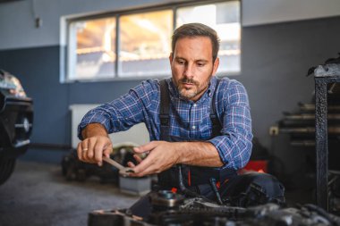 Bir araba tamircisi bir garajın içinde bir araba motorunu inceliyor ve bakım ya da onarım işine odaklanıyor. Görüntü detaylara ve teknik uzmanlığa dikkat çeker.