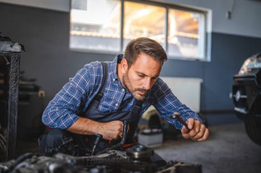 Bir araba tamircisi bir garajın içinde bir araba motorunu inceliyor ve bakım ya da onarım işine odaklanıyor. Görüntü detaylara ve teknik uzmanlığa dikkat çeker.