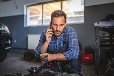 Araba tamircisi, garajda araç gereçle araba motorunu inceler ve cep telefonuyla konuşur.