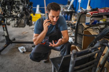 A car mechanic is multitasking, work on his laptop while engaged in a mobile phone call. He is in a garage setting, highlighting modern technology in auto repairs clipart