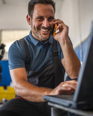 A car mechanic is multitasking, work on his laptop while engaged in a mobile phone call. He is in a garage setting, highlighting modern technology in auto repairs clipart