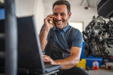Bir araba tamircisi birden fazla iş yapıyor, cep telefonuyla uğraşırken laptopuyla uğraşıyor. Otomobil tamirinde modern teknolojiyi ön plana çıkaran bir garaj ortamında.