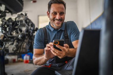 Mavi üniformalı neşeli bir araba tamircisi bir araba motorunun yanında oturur, cep telefonuyla parlak bir garaj ortamında meşgul olur, memnuniyeti ve modern teknolojiyi iş yerinde somutlaştırır.