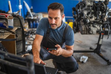 Odaklanmış bir araba tamircisi, araçlar ve parçalarla dolu modern bir garajda, bir araç motorunu incelemek için dizüstü bilgisayar ve cep telefonu kullanır.