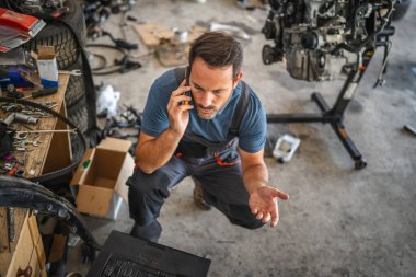 Atölyedeki bir tamirci cep telefonuyla konuşurken diz çöker, araba parçaları ve aletleri etrafa saçılmış, motor tamirini tartışır. Atölye kalabalık ve dağınık.