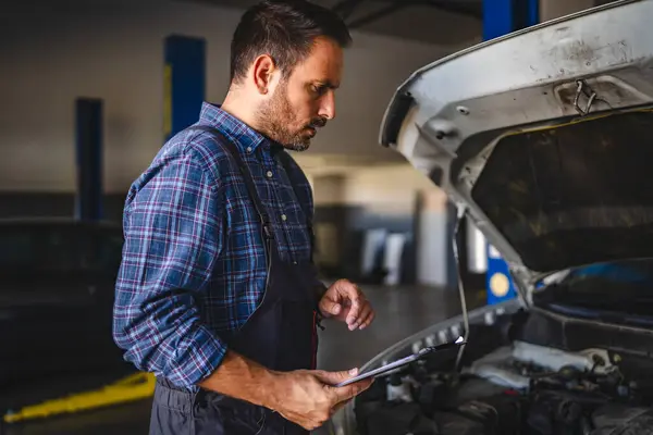 stock image A car mechanic in a car repair garage is examining the open engine compartment and reviewing diagnostics results on a clipboard, signifying diligent vehicle maintenance and service efficiency