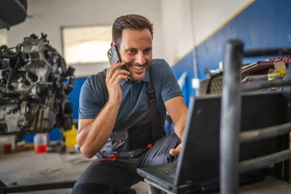 stock image A car mechanic is multitasking, work on his laptop while engaged in a mobile phone call. He is in a garage setting, highlighting modern technology in auto repairs