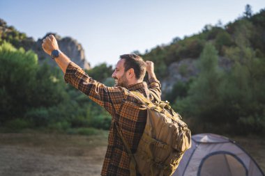 Bir kamp gezisinde geniş bir sırt çantası takmış, gün doğarken yemyeşil orman ve tepelerle çevrili bir çadırın yanında duran yetişkin bir adam yürüyüşçüsünün arkası.