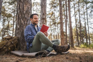 Yetişkin bir adam ağacın önünde oturup kitap okur.