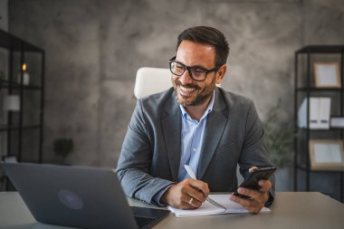 Kendine güvenen iş adamı masada oturur cep telefonu kullanır ve modern ofisteki deftere yazar.