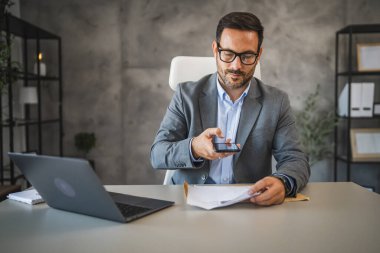 İşadamı ofisinde oturuyor ve cep telefonundan ödenmemiş faturalar alıyor.