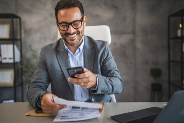 İşadamı ofisinde oturuyor ve cep telefonundan ödenmemiş faturalar alıyor.