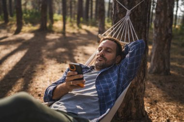 Yetişkin bir adam yatar ve hamakta cep telefonu kullanır.