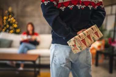 Noel süveteri giymiş bir erkek arkadaş, arkasında süslü bir Noel ağacı olan kanepesinde oturan kız arkadaşına hediyesini arkasına saklar.