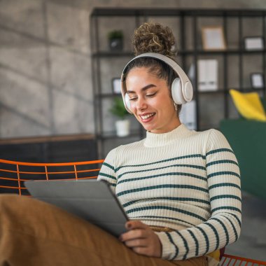 Genç bir kadın evde oturup dijital tablet ve kulaklık kullanarak film ya da dizi izlemek ya da mutlu bir gülümseme görüntüsü almak için iyi vakit geçirin.
