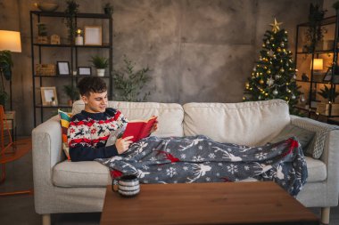 Genç adam kanepeye uzanıp şenlik battaniyesiyle kitap okuyor.