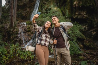 Yürüyüş yapan çift, şelalenin önünde kendi portresini ya da görüntüsünü çeker.