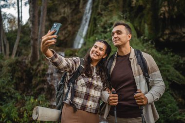 Yürüyüş yapan çift, şelalenin önünde kendi portresini ya da görüntüsünü çeker.