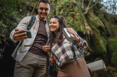 Yürüyüş yapan çift şelalenin önünde durup cep telefonu kullanıyor.