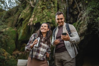 Yürüyüş yapan çift şelalenin önünde durup cep telefonu kullanıyor.