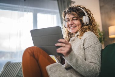 Kıvırcık saçlı beyaz bir kadın evde oturup dijital tablet ve kulaklık kullanarak film ya da dizi izliyor ya da mutlu bir gülümseme videosu çekiyor.