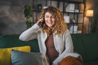 Evde kıvırcık saçlı beyaz bir kadın var. Cep telefonuyla telefon ediyor. Mutlu bir gülümseme.