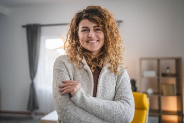 Evde kıvırcık saçlı, mutlu gülümseyen yetişkin beyaz kadın portresi.