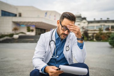 Endişeli doktor, panodan bilgi okuyup klinikteki merdivenlerde oturuyor.