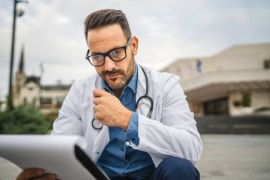 Endişeli doktor, panodan bilgi okuyup klinikteki merdivenlerde oturuyor.