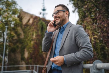 Gülümseyen yetişkin iş adamı terasta duruyor ve telefonla konuşuyor.