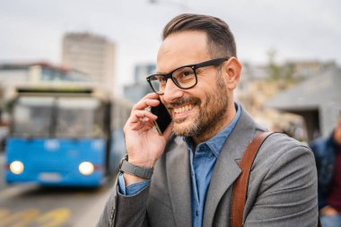 Yetişkin bir işadamının sokakta toplu taşımayı beklerken cep telefonuyla görüşmesi var.