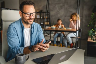 Baba evde çalışırken arka planda ailesini gezdirirken cep telefonu kullanıyor.