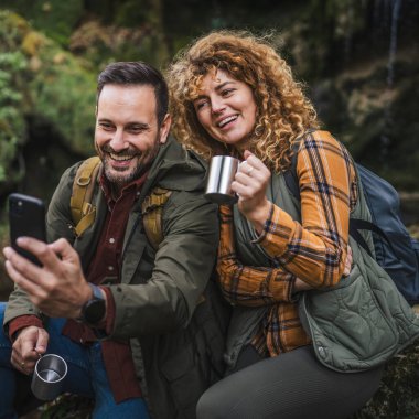 Karı koca yürüyüşçüleri mağaranın önünde oturup çay içerler ve görüntülü konuşma yaparlar ya da kendi portrelerini çekerler.